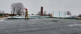 Zdjęcie przedstawia strażaka Państwowej Straży Pożarnej w umundurowaniu specjalnym z czerwonym hełmem na głowie. Strażak znajduje się na dachu budynku mieszkalnego. W tle widać uszkodzone pokrycie dachowe.