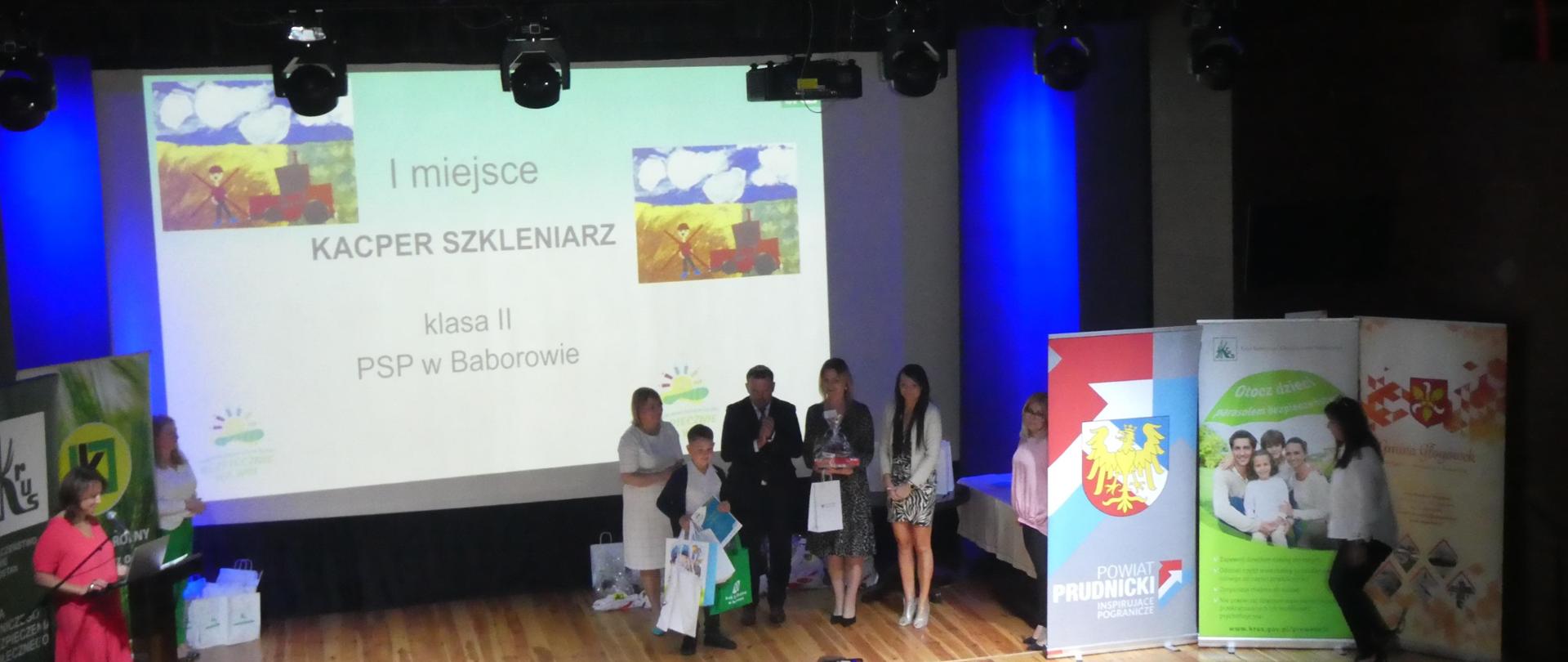 Na scenie laureat konkursu odbiera nagrody, za nim stoją trzy kobiety i mężczyzna, z tyłu ekran z prezentacją nazwiska laureata i zdjęciem jego pracy plastycznej.