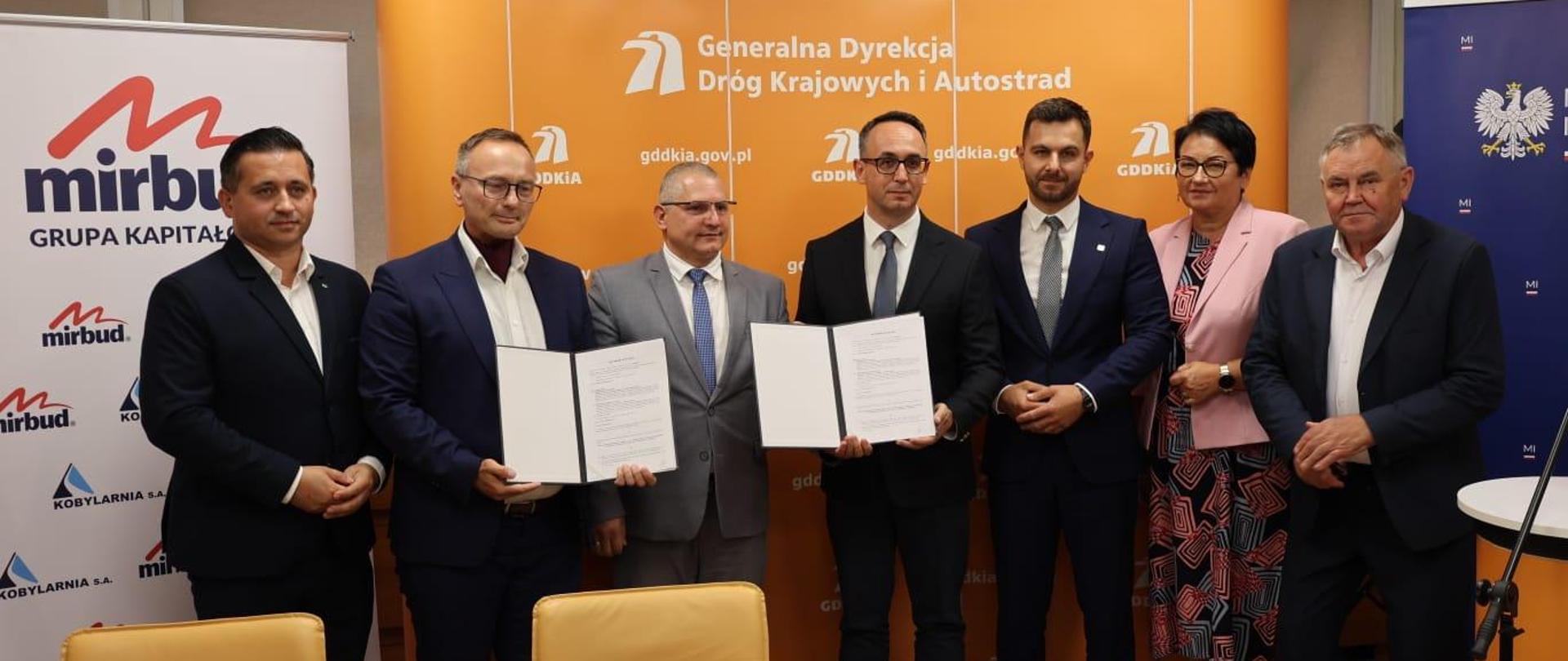 Wojewoda Opolski Monika Jurek, Minister Dariusz Klimczak oraz pozostali uczestnicy konferencji stoją i pozują do zdjęcia grupowego. Minister Dariusz Klimczak, Dyrektor Oddziału GDDKiA w Opolu Rafał Pydych oraz Wiceprezes Kobylarnia S.A. Michał Niemyt trzymają w rękach podpisane umowy. 