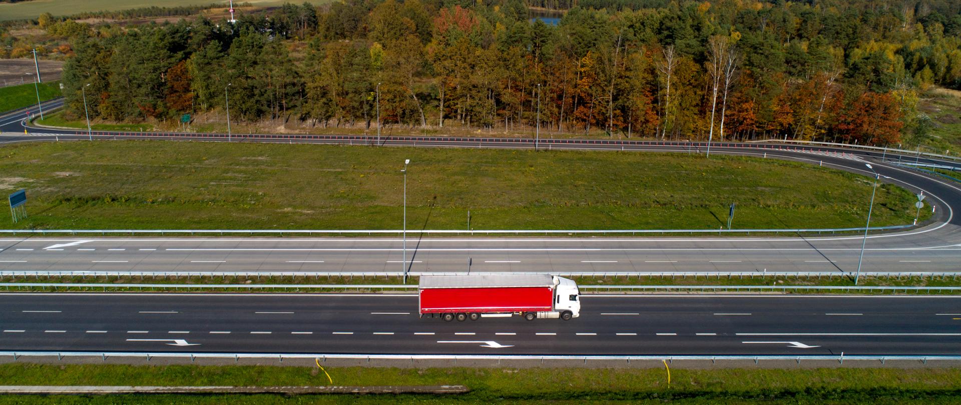 Droga dwujezdniowa - autostrada A18 w otoczeniu lasów. Ujęcie drogi w poziomie, sceneria jesienna. Na drodze samochód ciężarowy z czerwoną naczepą.