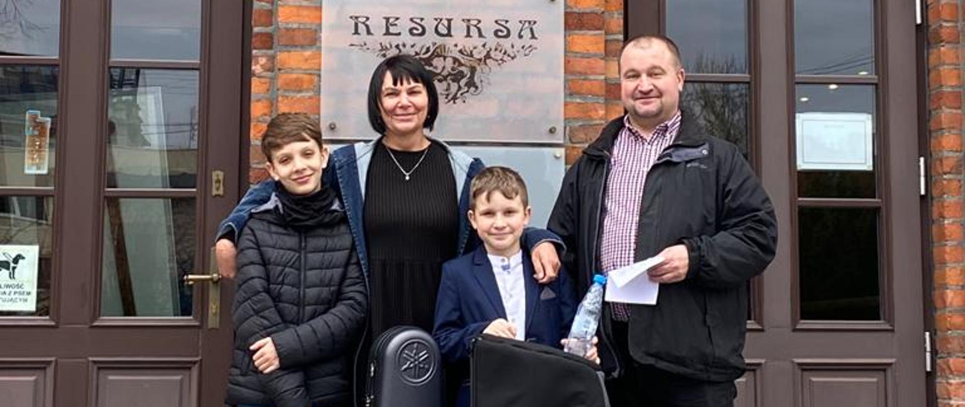 Zdjęcie przedstawia uczniów: Tomasz Weinkopf i Karol Ludwig, nauczyciela: Andrzej Weinkopf i akompaniator" Mirosława Ignac stojących przed budynkiem Resursy w Żyrardowie
