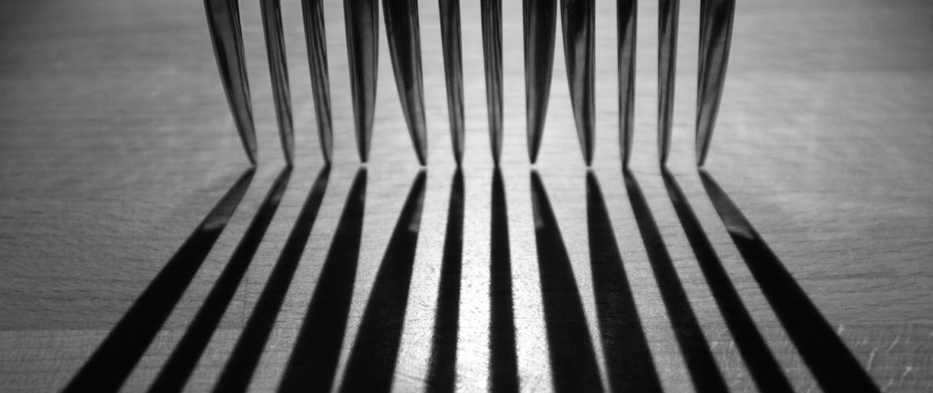 Zdjęcie jest w kolorystyce czarno-białej. Zdjęcie przedstawia zęby trzech widelców postawione obok siebie, które rzucają cień na podłoże.