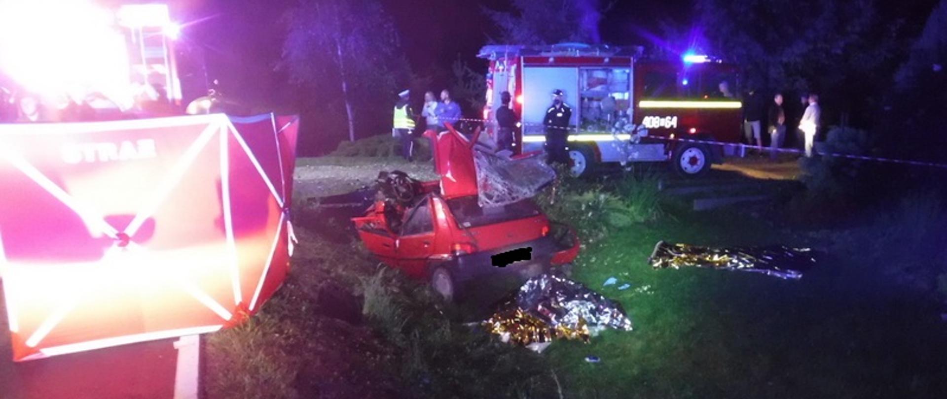 Zdjęcie główne przedstawia rozbity samochód osobowy w przydrożnym rowie. W pobliżu samochodu znajdują się zwłoki dwóch osób przykryte przez strażaków folią NRC. W pobliżu rozbitego samochodu stoją dwa samochody ratowniczo- gaśnicze z włączonymi lampami błyskowymi. Na miejscu zdarzenia pracują strażacy z JRG Bochnia oraz OSP Baczków i OSP Dziewin. 