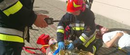 Trzech strażaków udzielających pomocy medycznej dwóm poszkodowanym uczniom na chodniku przed szkołą