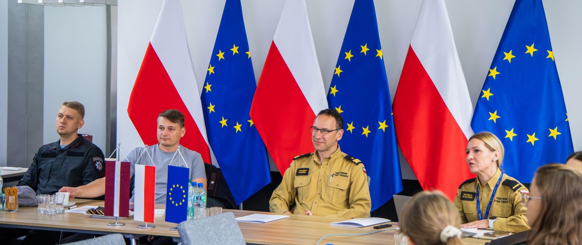 Uczestnicy spotkania siedzą przy stole w sali konferencyjnej. W tle stoją flagi Polski i Unii Europejskiej. Na środku stołu umieszczono miniaturowe flagi Polski, Łotwy i UE.