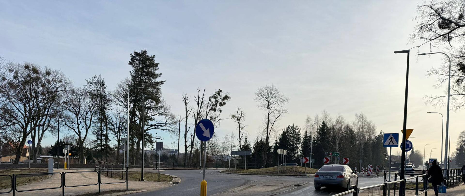 Ulica w mieście z wysepką rozdzielającą pasy ruchu. Na pierwszym planie widoczne są metalowe barierki oddzielające chodnik od jezdni. Na środku znajduje się znak nakazu jazy z prawej strony zamocowany na słupku z żółtą podstawą. W tle widoczny jest samochód osobowy poruszający się po drodze, a także inne pojazdy i piesi. Po prawej stronie znajduje się przejście dla pieszych oznaczone znakiem drogowym. Krajobraz zawiera bezlistne drzewa, co sugeruje porę zimową lub jesień. 