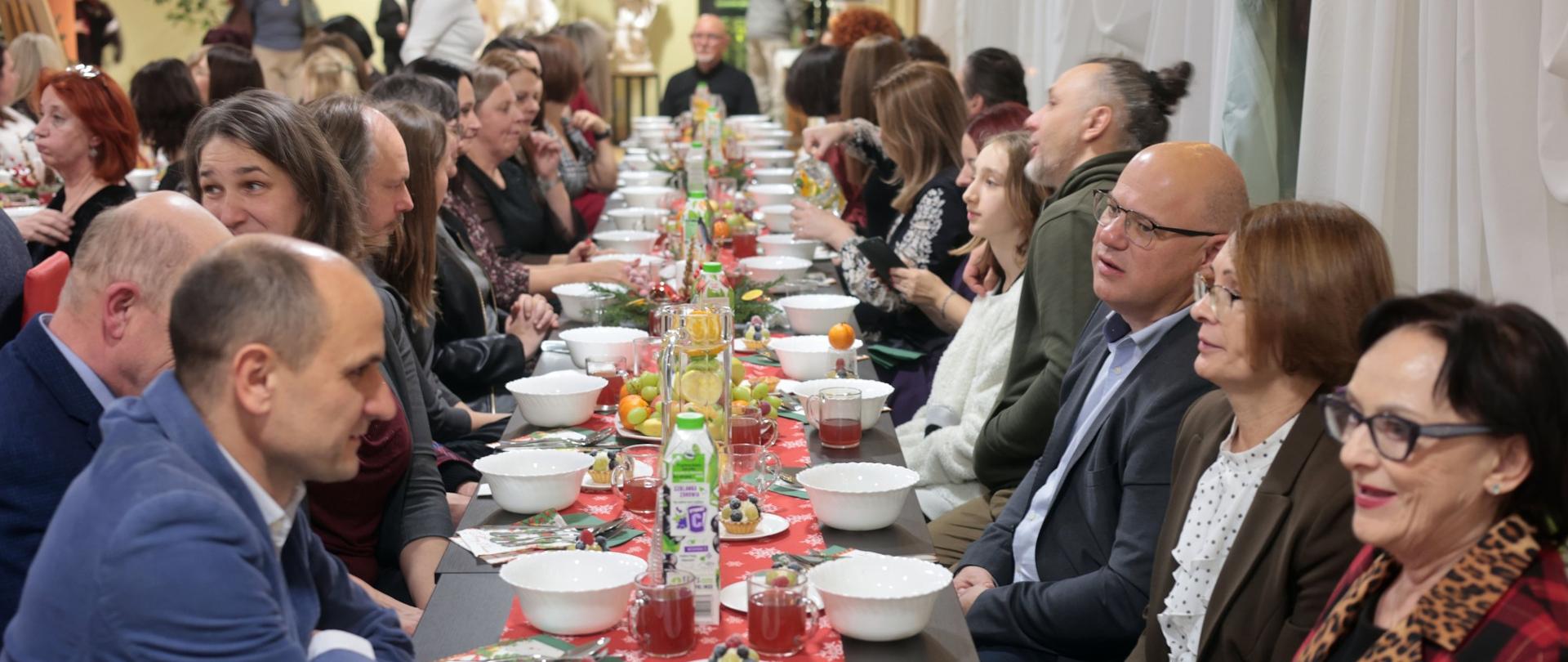 Zdjęcie przedstawia długie, odświętnie udekorowane stoły ustawione w sali, na których znajdują się nakrycia do wspólnego posiłku. Na stołach zastawa: białe miski, sztućce, kubki z czerwonymi serwetkami, dzbanki z napojami.
Dekoracje: świąteczne elementy, takie jak stroiki, bombki, świeczki, oraz owoce (mandarynki).
Tło: choinka ozdobiona światełkami, świąteczne dekoracje na ścianach i oknach, co podkreśla bożonarodzeniowy klimat.
Całość wygląda na wigilię organizowaną w gronie pracowników, rodziny lub społeczności, w eleganckiej, świątecznej atmosferze.