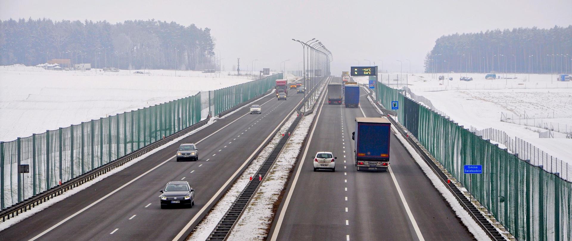 Dwujezdniowa droga szybkiego ruchu. Duży ruch pojazdów osobowych i ciężarowych. Po obu stronach drogi rozwieszone zielone siatki. Na pasie rozdzielającym jezdnie i poza drogą leży śnieg.