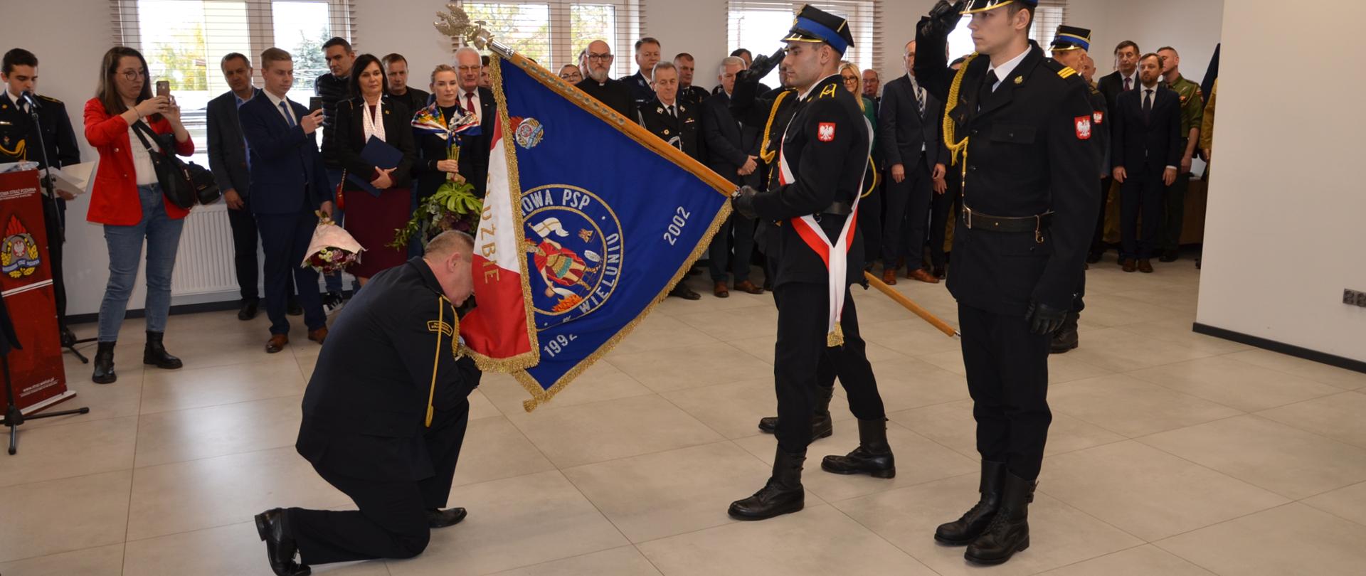 Scena ceremonii, podczas której strażak w ciemnym mundurze klęczy i całuje sztandar Komendy Powiatowej PSP w Wieluniu. Dwóch innych strażaków w mundurach i czapkach salutuje obok sztandaru. W tle stoi większa grupa osób, w tym strażacy i cywile.
