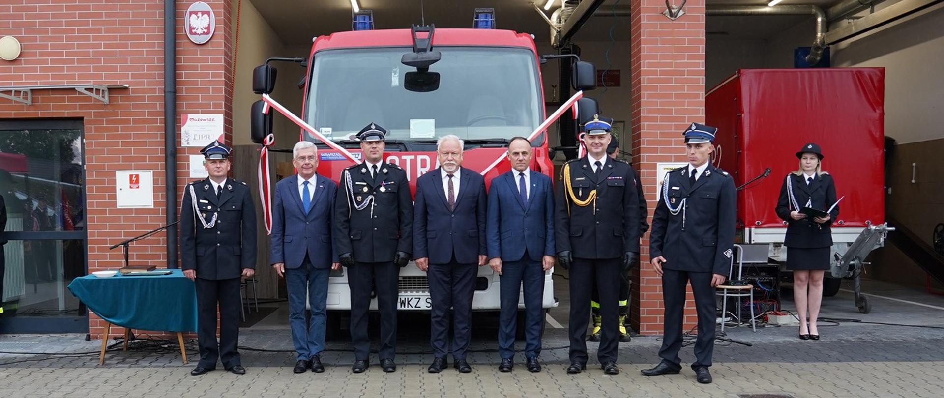 zaproszeni gości stoją przed samochodem pożarniczym który stoi w bramie remizy OSP