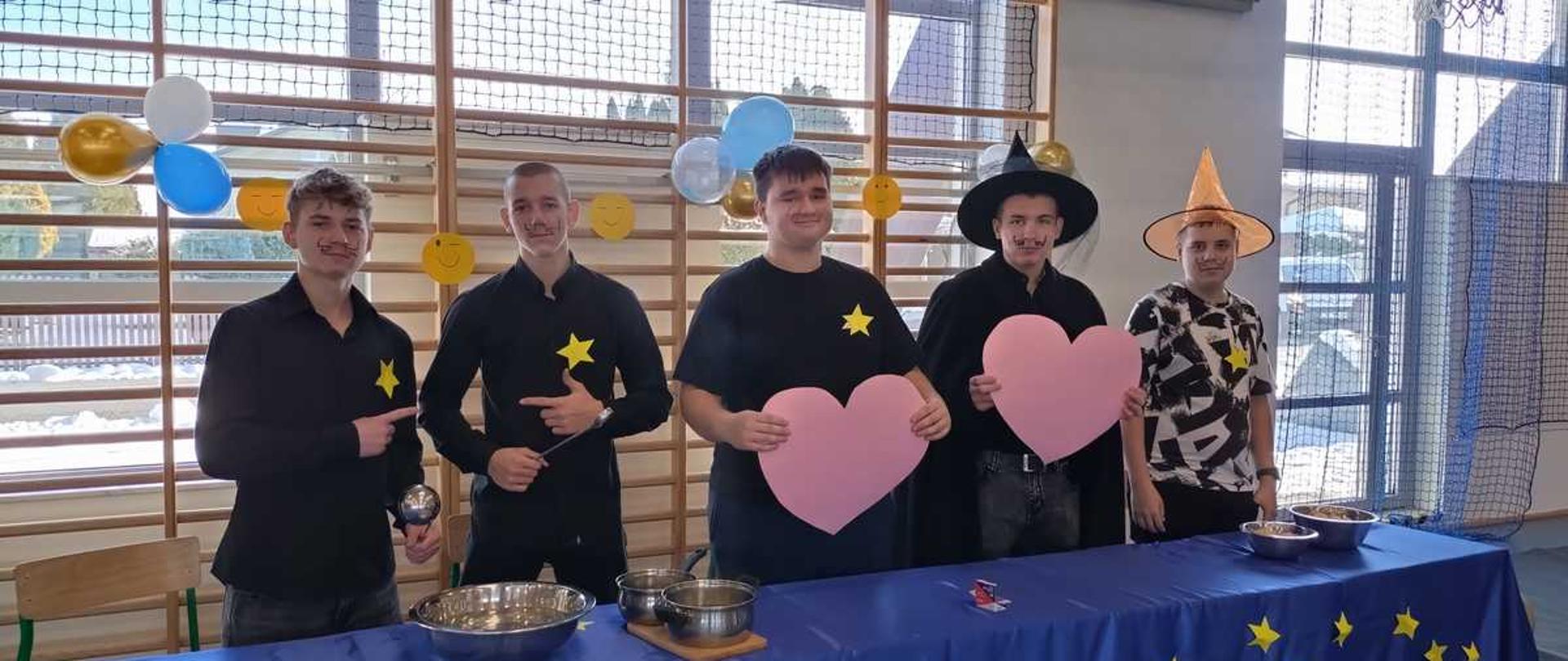 Zdjęcie przedstawia pięciu młodych mężczyzn, prawdopodobnie uczniów szkoły, stojących za stołem z niebieskim obrusem, na którym widnieją żółte gwiazdki (symbol Unii Europejskiej). W tle widać salę gimnastyczną.
Opis szczegółowy:
Od lewej:
Pierwszy chłopak (po lewej stronie) ubrany jest w czarną koszulkę z żółtą gwiazdą Dawida, dżinsy i wskazuje lewą ręką na rekwizyt w kształcie kryształowej kuli trzymany przez kolejnego chłopaka.
Drugi chłopak (drugi od lewej) ma na sobie czarną koszulkę z żółtą gwiazdą i dżinsy, a w prawej ręce trzyma wspomnianą kulę.
Trzeci chłopak (środek) ubrany jest w czarną koszulkę z żółtą gwiazdą i trzyma duży, różowy rekwizyt w kształcie serca.
Czwarty chłopak (drugi od prawej) ma na sobie czarną pelerynę, czarny kapelusz czarodzieja i trzyma drugi duży, różowy rekwizyt w kształcie serca.
Piąty chłopak (po prawej) ubrany jest w kolorową koszulkę z nadrukami i ma na głowie spiczasty, pomarańczowy kapelusz czarodzieja.
Na stole znajdują się miski i garnki. W tle wiszą białe, złote i niebieskie balony. Całość stylizacji i rekwizytów sugeruje, że jest to szkolne przedstawienie, możliwe, że związane z magią, Halloween lub andrzejkami. Niektóre wyniki wyszukiwania sugerują, że podobne wydarzenia mogą być organizowane w ramach obchodów Dni Europy, co pasowałoby do symboli gwiazd na obrusie.