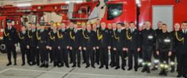 strażacy uczestnicy uroczystości pozują do zdjęcia, stojąc w grupie na tle samochodów pożarniczych