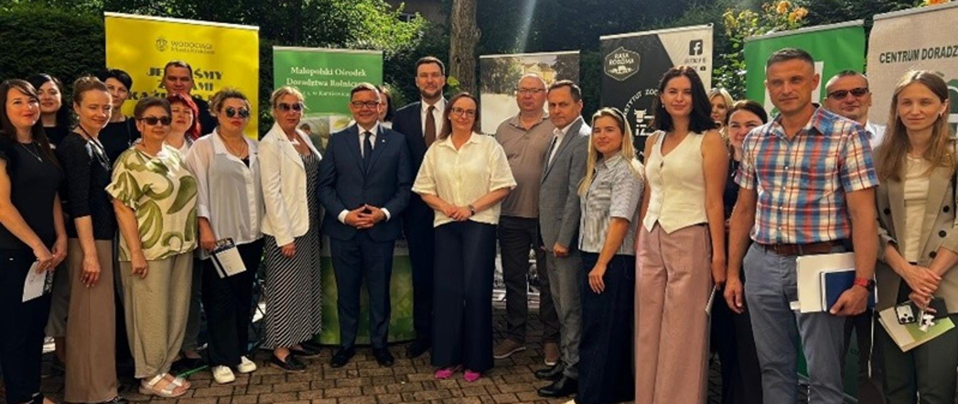 Grupa osób stojących w półkolu na tle kolorowych banerów reklamowych instytucji rolniczych, m.in. KRUS. Po środku stoją elegancko ubrani dwaj mężczyźni w białych koszulach, krawatach i ciemnych marynarkach, którzy reprezentują Ministerstwo Rolnictwa i Rozwoju Wsi oraz Instytut Zootechniki – Państwowy Instytut Badawczy. Wszystkie osoby lekko się uśmiechają, pozując do zdjęcia. Za nimi znajdują się stoliki informacyjno-prewencyjne z ulotkami informacyjnymi i gadżetami prewencyjnymi. 
