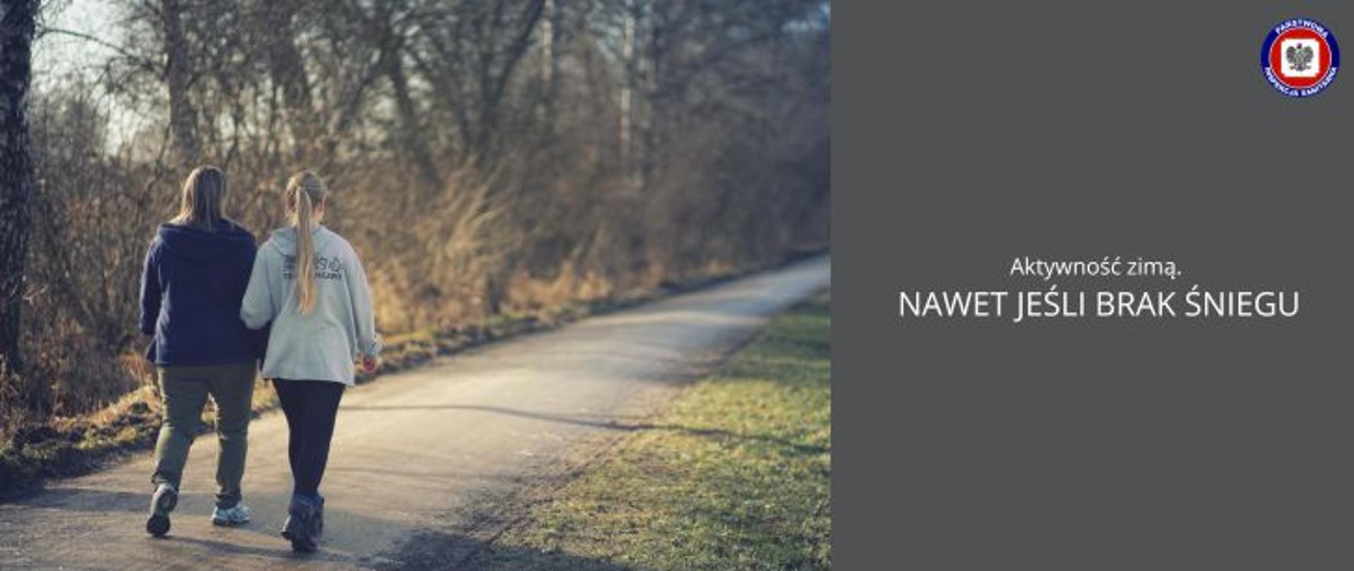 Po leśnej drodze spacerują 2 dziewczyny ubrane na sportowo. Po prawej stronie na grafitowym tle jasny napis Aktywność zimą. Nawet jeśli brak śniegu. W prawym górnym rogu logo Państwowej Inspekcji Sanitarnej.