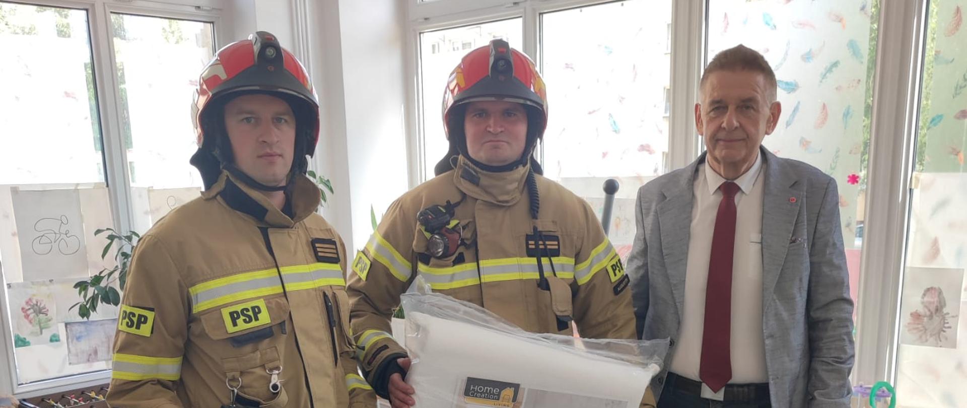 Na zdjęciu widać strażaków przekazujących paczkę przedstawicielowi Hospicjum w Augustowie