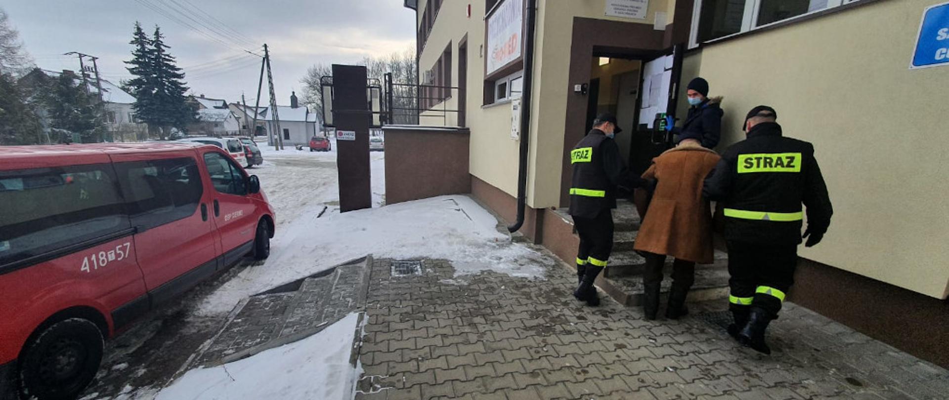 Na zdjęciu przedstawiony jest po lewej stronie samochód operacyjny OSP. Po prawej stronie ukazani są dwaj strażacy w umundurowaniu koszarowym z założonymi maseczkami ochronnymi na twarz. Pomagają oni wejść po schodach do budynku Ośrodka Zdrowia starszemu mężczyźnie, który udaje się w ich asyście na szczepienie przeciwko Covid- 19.