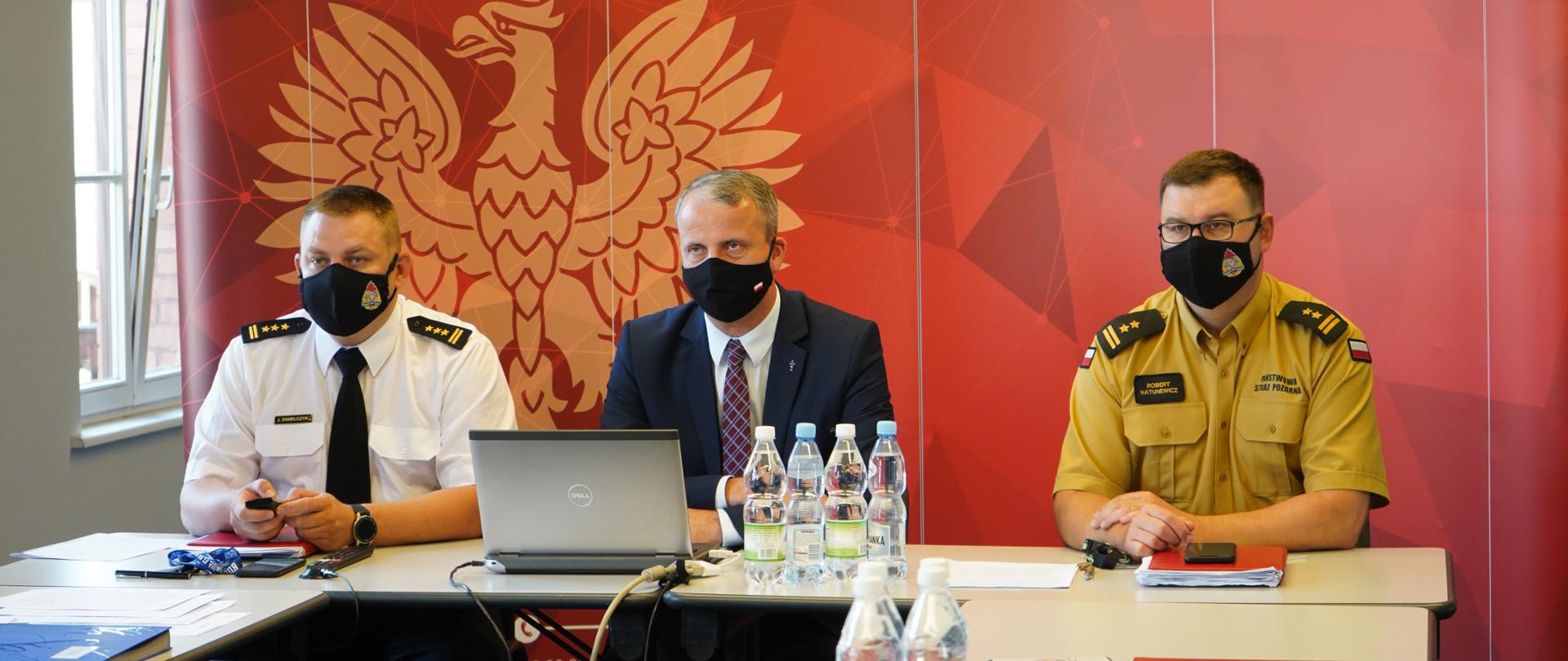 trzech mężczyzn siedzi orzy stole w maseczkach ochronnych, porzed nimi komputer oraz butelki z wodą, w tle baner z orłem