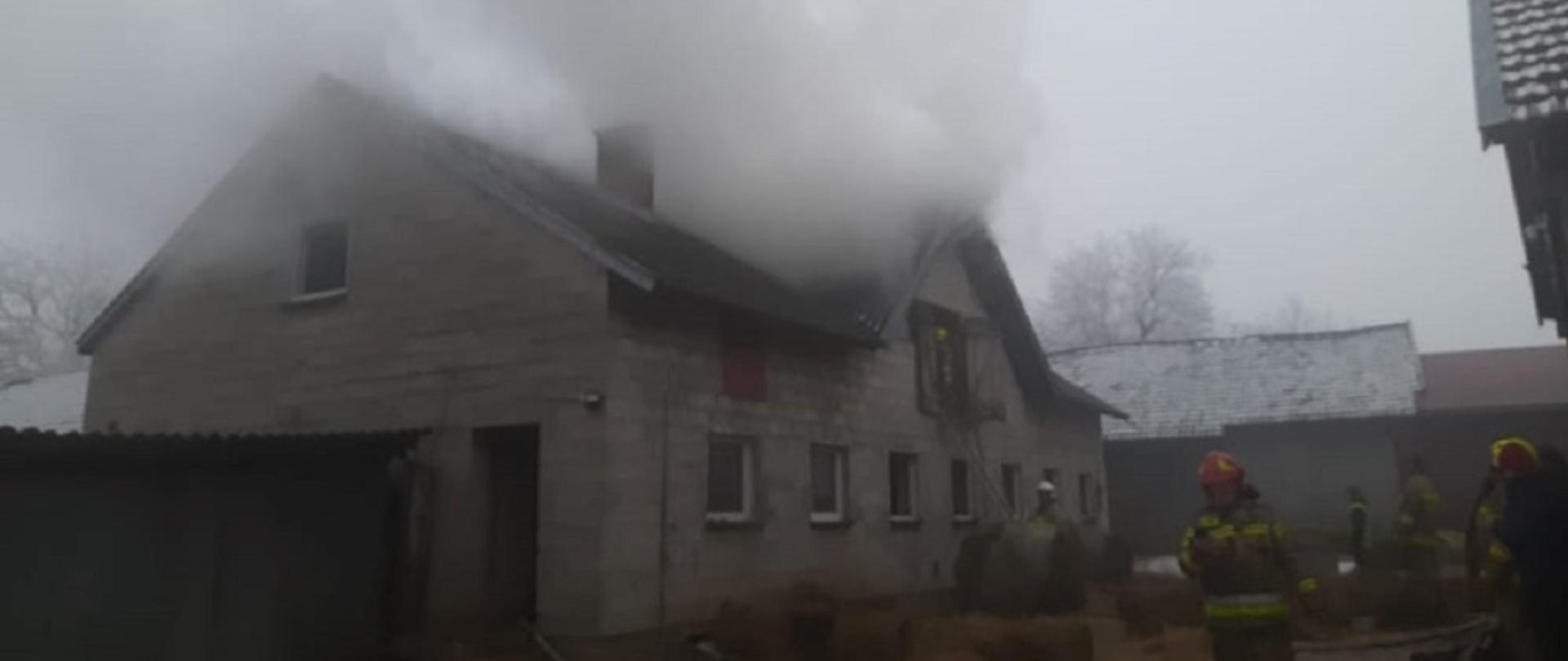 Na zdjęciu zadymiona część więźby dachowej budynku gospodarczego, przed budynkiem strażacy prowadzący działania gaśnicze.