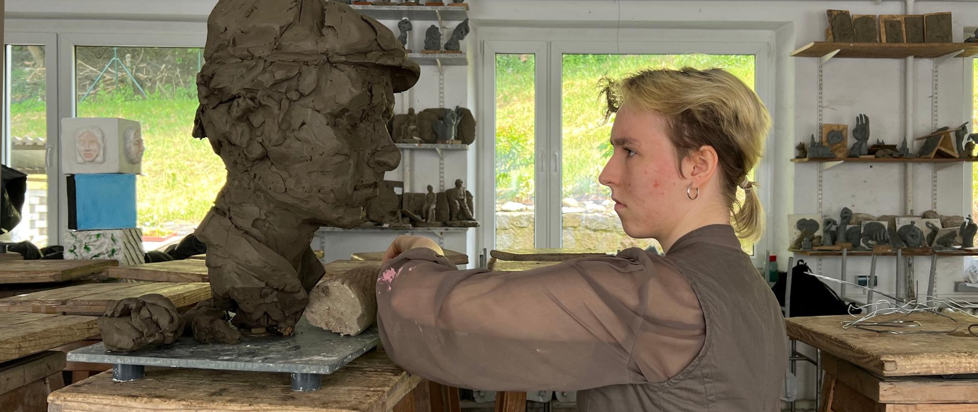 Zdjęcie wykonane w pracowni rzeźbiarskiej. Młoda rzeźbiarka pracuje w glinie nad rzeźbą głowy mężczyzny. W tle kawalety, pułki na ścianie i na nich drobne prace rzeźbiarskie. 