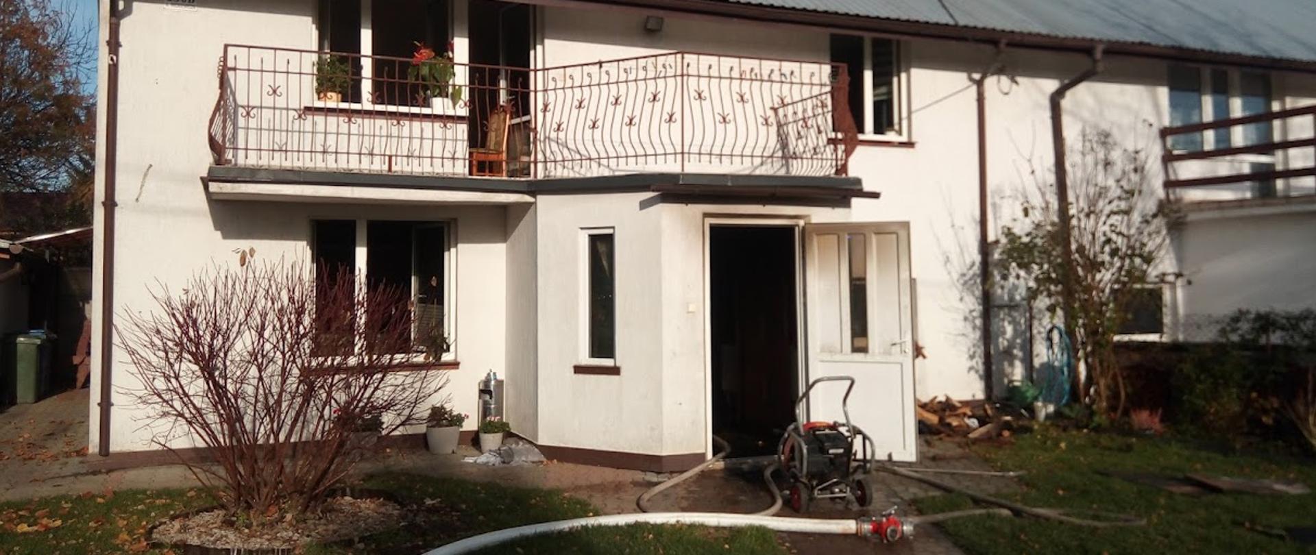Zdjęcie przedstawia biały piętrowy budynek mieszkalny z otwartymi oknami i okopconymi drzwiami wejściowym, przed budynkiem leży biały wąż gaśniczy 