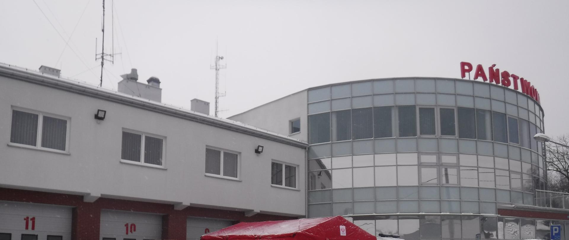 Czerwony namiot do którego jest ustawiona kolejka ludzi , którzy stoją w odstępach zachowując dystans około 1m. Są to nauczyciele ze szkół z terenu powiatu Bocheńskiego, którzy oczekują na pobranie materiału do wymazu na Covid. Koło namiotu po lewej stronie stoi karetka pogotowia w której personel medyczny zajmuje się pobieraniem próbek. Za namiotem, który jest rozstawiony przed komendą na parkingu straży pożarnej w Bochni zaparkowany jest bus ochotniczej straży pożarnej w Rzezawie. 