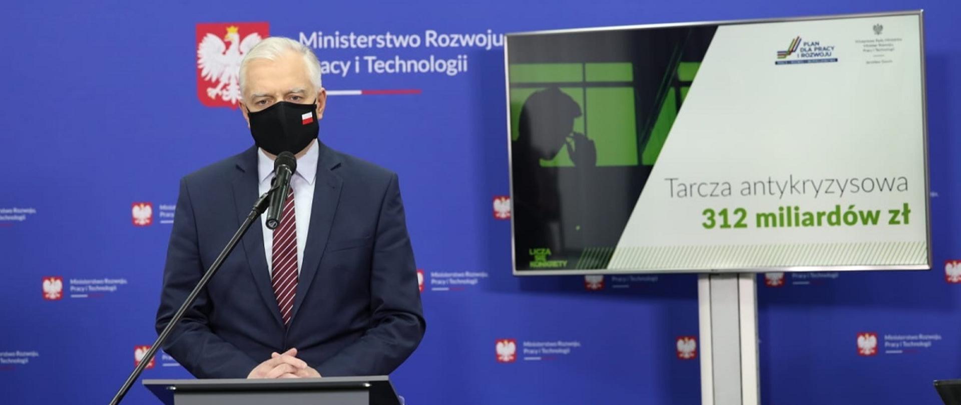 Wicepremier i minister rozwoju, pracy i technologii Jarosław Gowin stojący w maseczce przed mównicą, po jego prawej stronie ekran z napisem: :"Tarcza antykryzysowa 312 miliardów"
