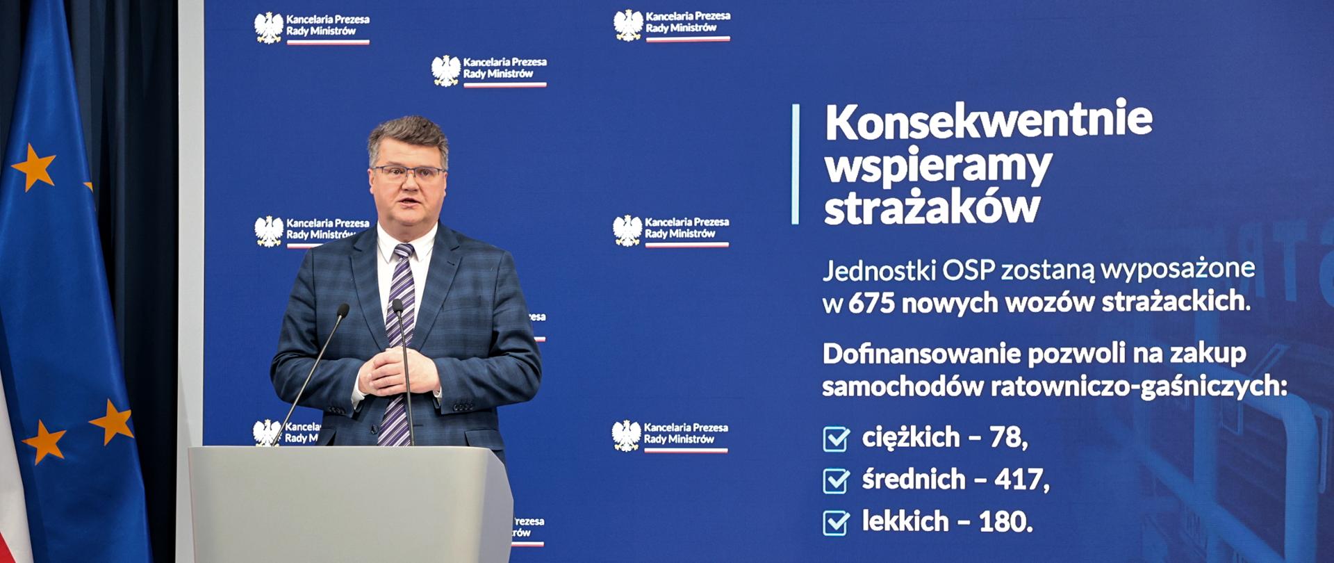 Zdjęcie przedstawia Wiceministra Macieja Wąsika stojącego przy mównicy podczas konferencji prasowej dotyczącej zakupu 675 samochodów strażackich dla jednostek OSP z terenu kraju. W tle slajd z Kancelarii Prezesa Rady Ministrów.