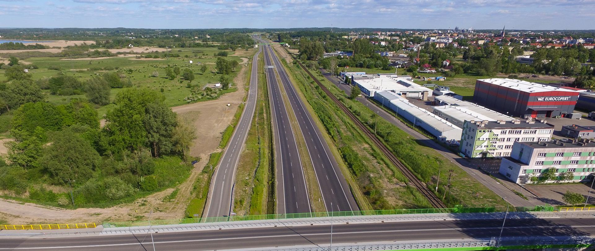 Droga ekspresowa S11. Dwujezdniowa droga ekspresowa po dwa pasy ruchu w obu kierunkach. Po prawej stronie widać zabudowania Szczecinka. Wzdłuż drogi biegną tory kolejowe.