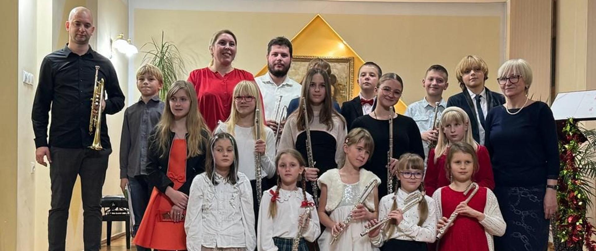 Grupa uczniów, którzy trzymają instrumenty wraz z nauczycielami P. W. Lizurejem, i P. B Adamską pozują do zdjęcia grupowego w auli PSM w Kłodzku podczas koncertu uczniów klasy fletu, trąbki i fortepianu - 01 grudnia 2023