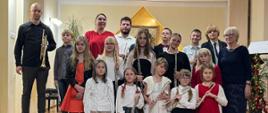 Grupa uczniów, którzy trzymają instrumenty wraz z nauczycielami P. W. Lizurejem, i P. B Adamską pozują do zdjęcia grupowego w auli PSM w Kłodzku podczas koncertu uczniów klasy fletu, trąbki i fortepianu - 01 grudnia 2023
