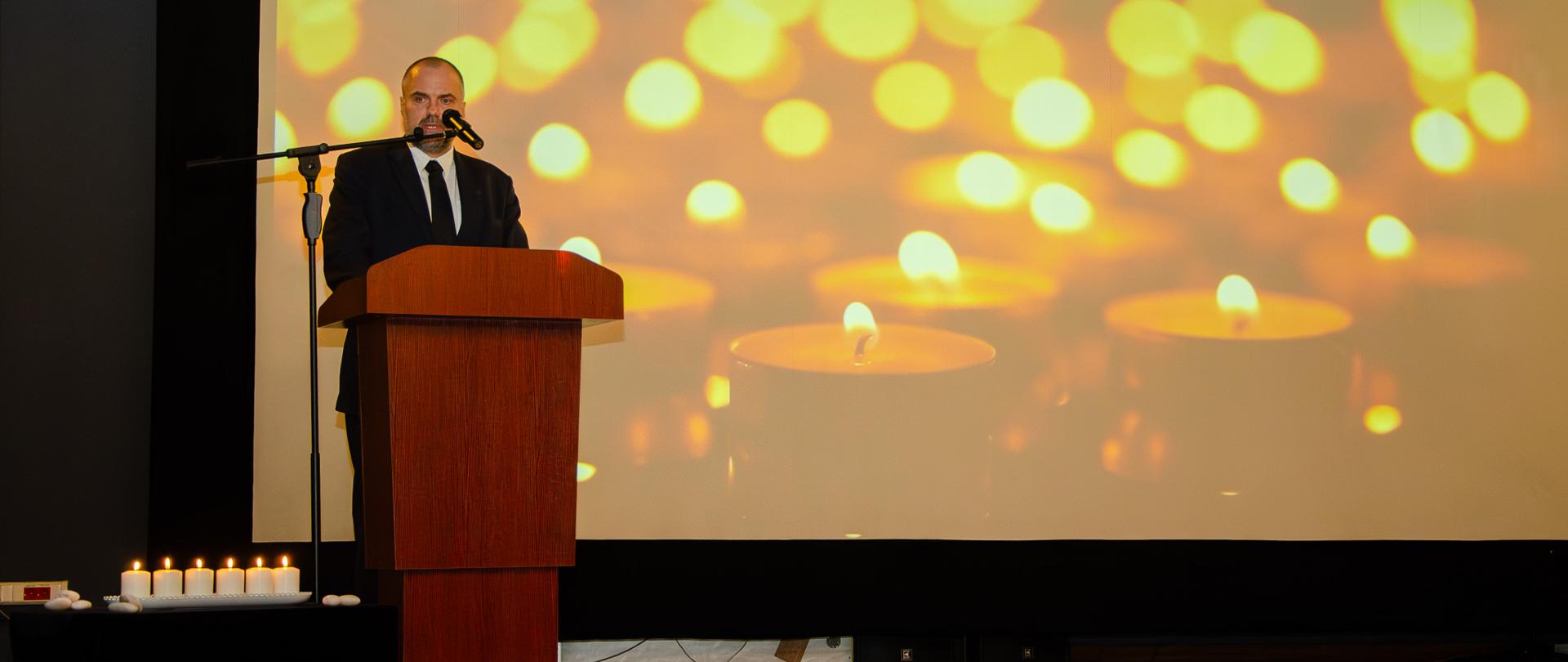 Ambasador Jakub Kumoch na scenie podczas przemówienia na tle ekranu ze świecami