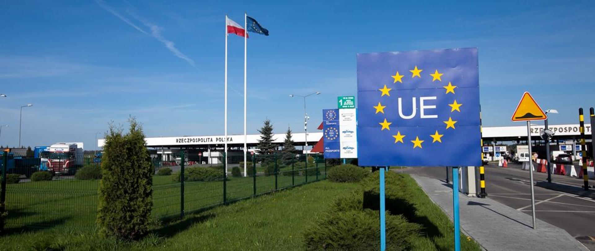 Przejście graniczne z widocznym znakiem „UE”, flagami Polski i Unii Europejskiej oraz budynkami odprawy granicznej w tle. Do przejścia prowadzi droga z wydzielonymi pasami ruchu.