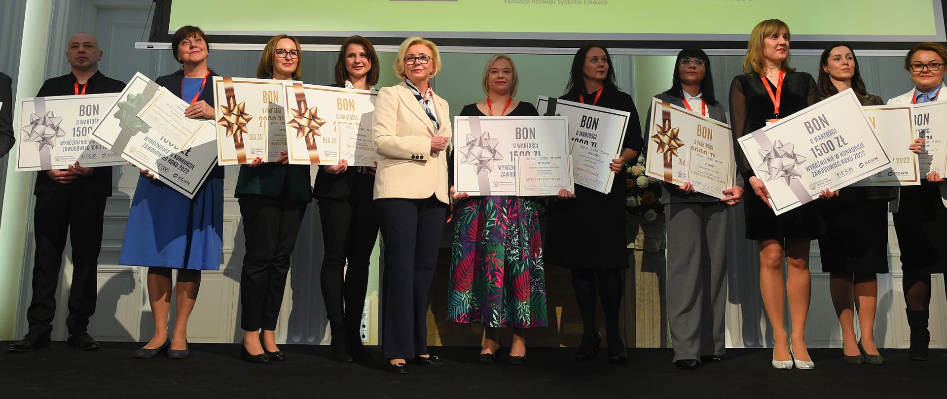 Pod wielkim ekranem stoi grupa ludzi trzymających duże kartki z napisem BON, przed nimi wiceminister Machałek w białej marynarce.