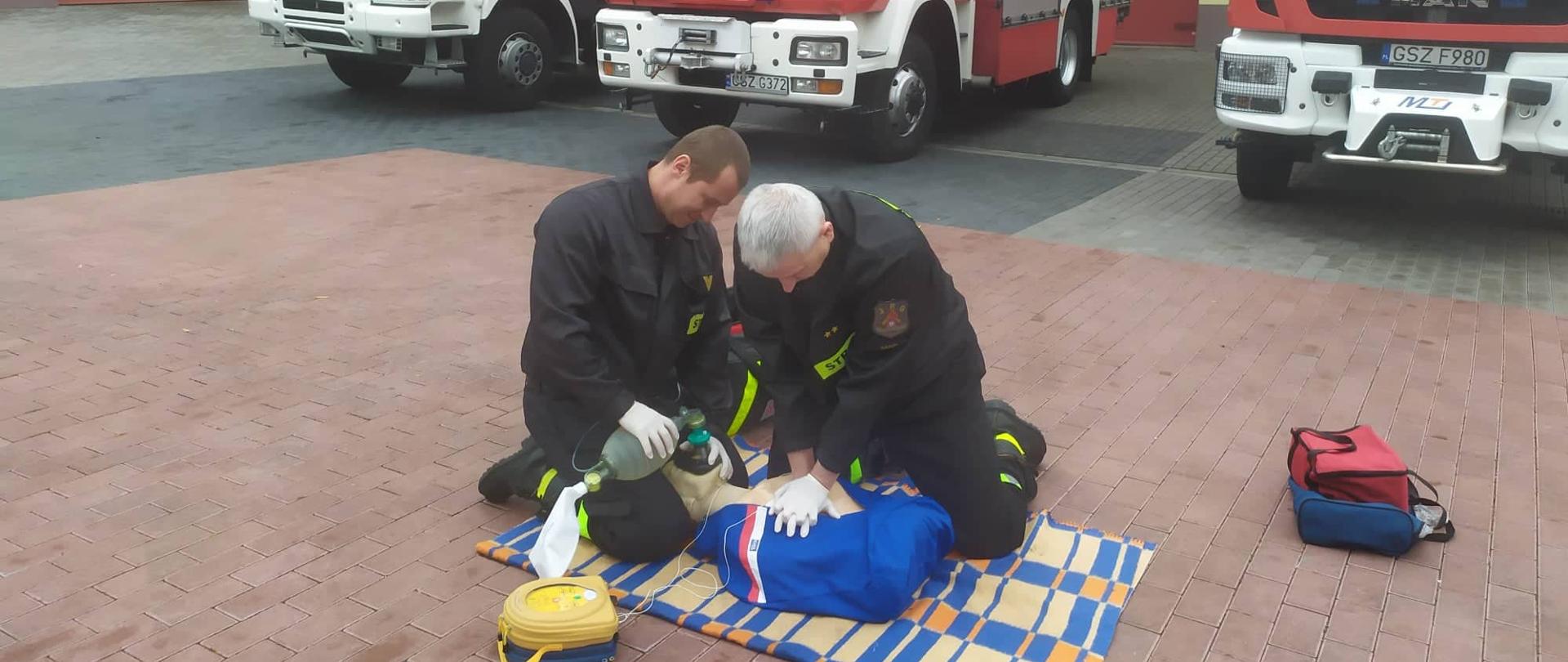 na placu brukowanym, dwóch strażaków przywraca czynności życiowe w formie ćwiczebnej na manekinie. W tle widać pojazdy pożarnicze. 