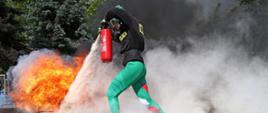 Zawodnik w zielonym stroju gasi palący się palnik przy użyciu gaśnicy proszkowej. Duża ilość dymu i intensywny ogień kontrastują z szybkim i zdecydowanym ruchem strażaka w masce ochronnej.