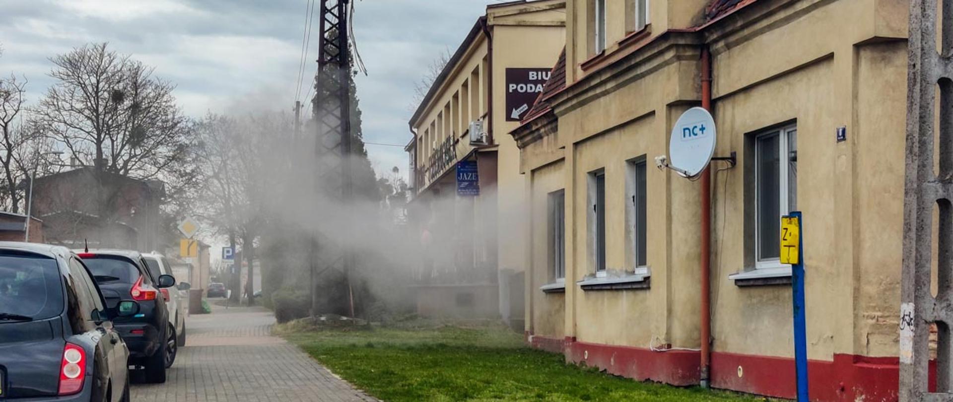 Na zdjęciu budynek wielorodzinny gdzie z jednego okna wydobywa się dym obok stoją przy chodniku samochody osobowe na budynku jest talerz od telewizji satelitarnej