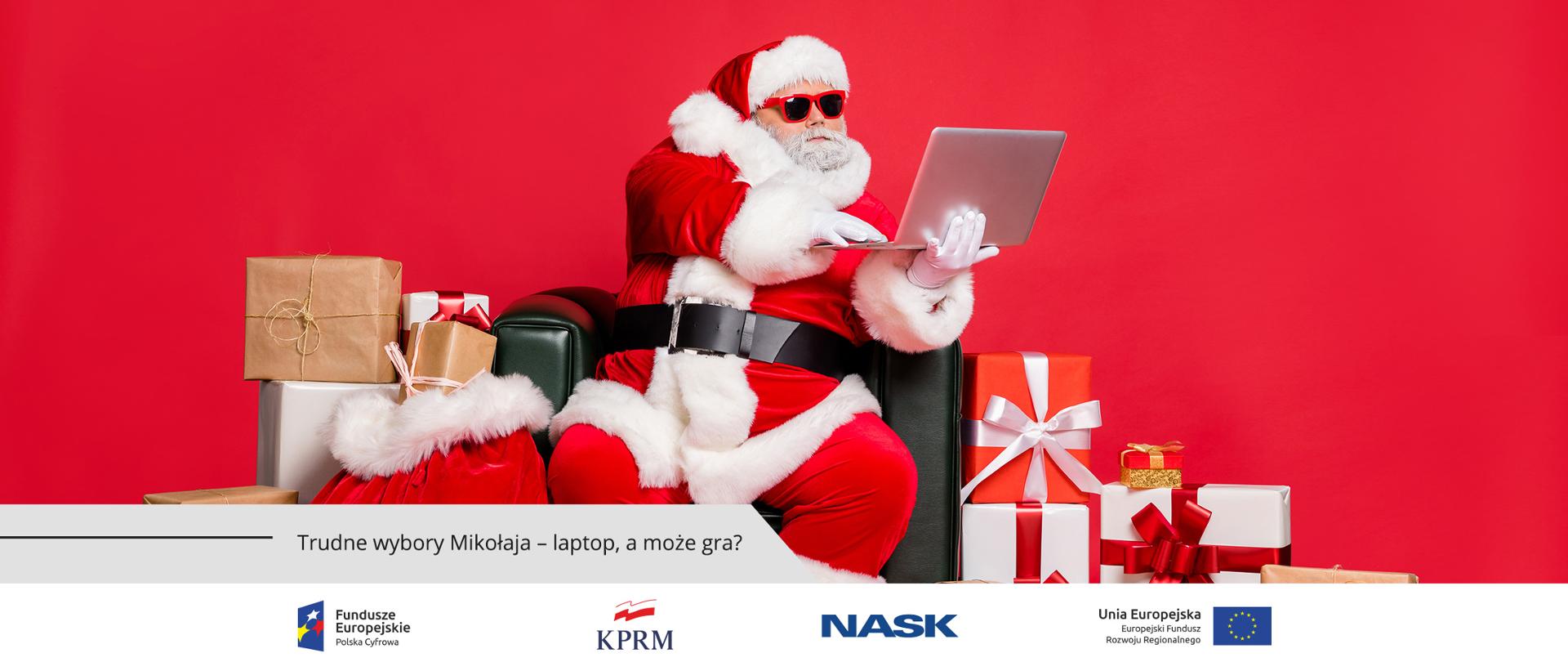 Mężczyzna w stroju św. Mikołaja, siedzi na czarnym, skórzanym fotelu, na oczach ma okulary przeciwsłoneczne, a w rękach trzyma laptopa. Wokół niego leżą zapakowane prezenty.