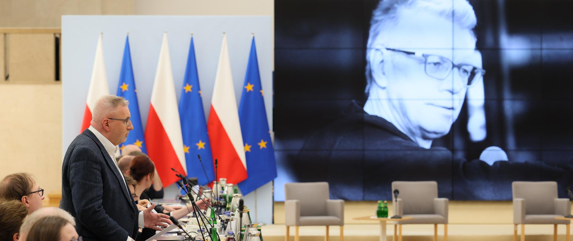 Szeroki kadr. Na zdjęciu po lewej stronie widoczni członkowie Rady przy stole konferencyjnym. Pan Krzysztof Balon stojąc wspomina śp. Pana Cezarego Miżejewskiego. Po prawej stronie, na dużym ekranie, jest wyświetlone czarno-białe zdjęcie Pana Cezarego Miżejewskiego. Z lewej strony ekranu są ustawione flagi Polski i Unii Europejskiej.