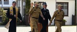 Dwóch strażaków w mundurach koloru musztardowego kapelan oraz strażak ochotniczej straży pożarnej idą obok siebie za nimi na baczność stoi funkcjonariusz Państwowej Straży Pożarnej.
