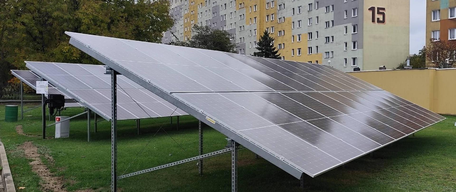 Na zdjęciu przedstawiono trzy stanowiska z panelami fotowoltaicznymi. Stanowiska o metalowej konstrukcji ustawiono na trawniku znajdującym się na placu wewnętrznym komendy. W tle widoczne budynki wielorodzinne.