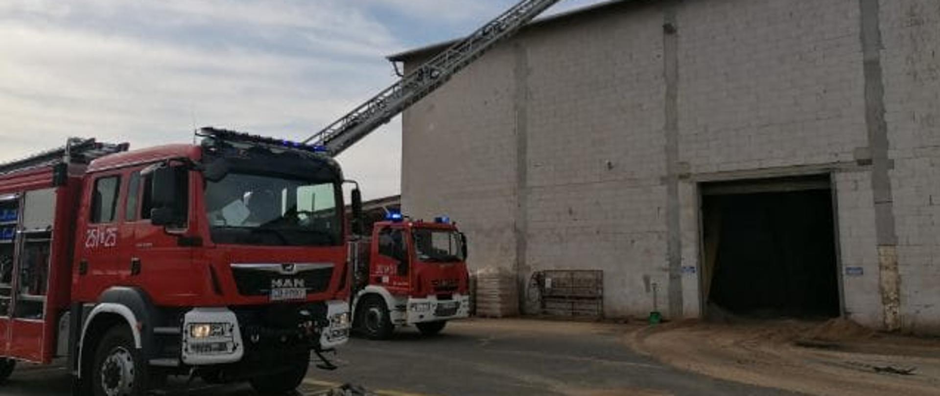 W lewej części zdjęcia przód pojazdu strażackiego ciężarowego koloru czerwonego marki MAN należącego do Jednostki Ratowniczo-Gaśniczej Szkoły Podoficerskiej PSP w Bydgoszczy, w tle przód pojazdu pożarniczego drabiny mechanicznej z JRG1 w Bydgoszczy, pojazdy stoją przy hali, gdzie przebiega pożar. Konstrukcja hali z zewnątrz w stanie surowym z pustaków gazobetonowych.