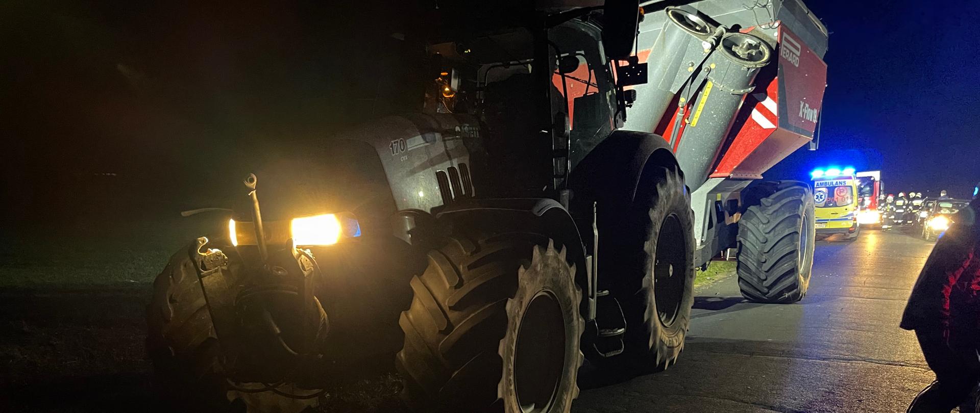 Na zdjęciu widać ciągnik rolniczy z przyczepą, który brał udział w zdarzeniu. W tle za ciągnikiem widać karetkę pogotowia oraz samochód pożarniczy. Obok pojazdów stoją strażacy. Z uwagi na ciemności strażacy oświetlają teren działań