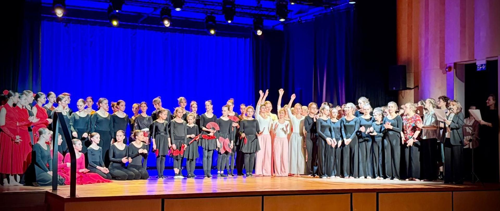 Zdjęcie kolorowe. Grupa tancerek i tancerzy na drewnianej scenie w sali widowiskowej. Osoby ubrane w czarne, czerwone i białe stroje baletowe lub kostiumy sceniczne. Niektóre tancerki trzymają czerwone wachlarze. Część grupy stoi w rzędach, kilka osób siedzi na deskach sceny po lewej stronie, a inne unoszą ręce w górę. Po prawej stronie stoją osoby w strojach codziennych, trzymające kartki papieru. Tło stanowi granatowa kurtyna oświetlona jasnoniebieskim światłem z reflektorów zamontowanych na czarnym stelażu pod sufitem. W dolnej części ujęcia widoczne oparcia foteli na widowni oraz zarysy głów siedzących osób. Sala ma jasne, drewniane ściany boczne. Panuje podniosła atmosfera po występie