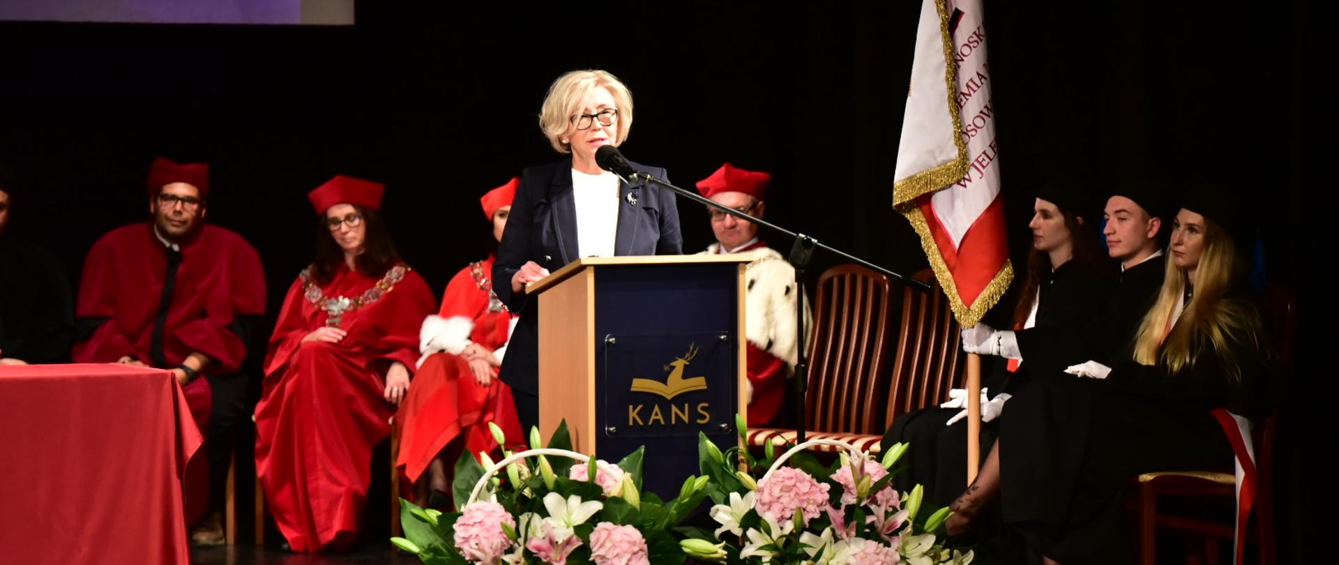 Wiceminister Machałek przemawia przy mikrofonie, przed nią stoją kosze kwiatów, za nią siedzą władze uczelni, obok studenci ze sztandarem