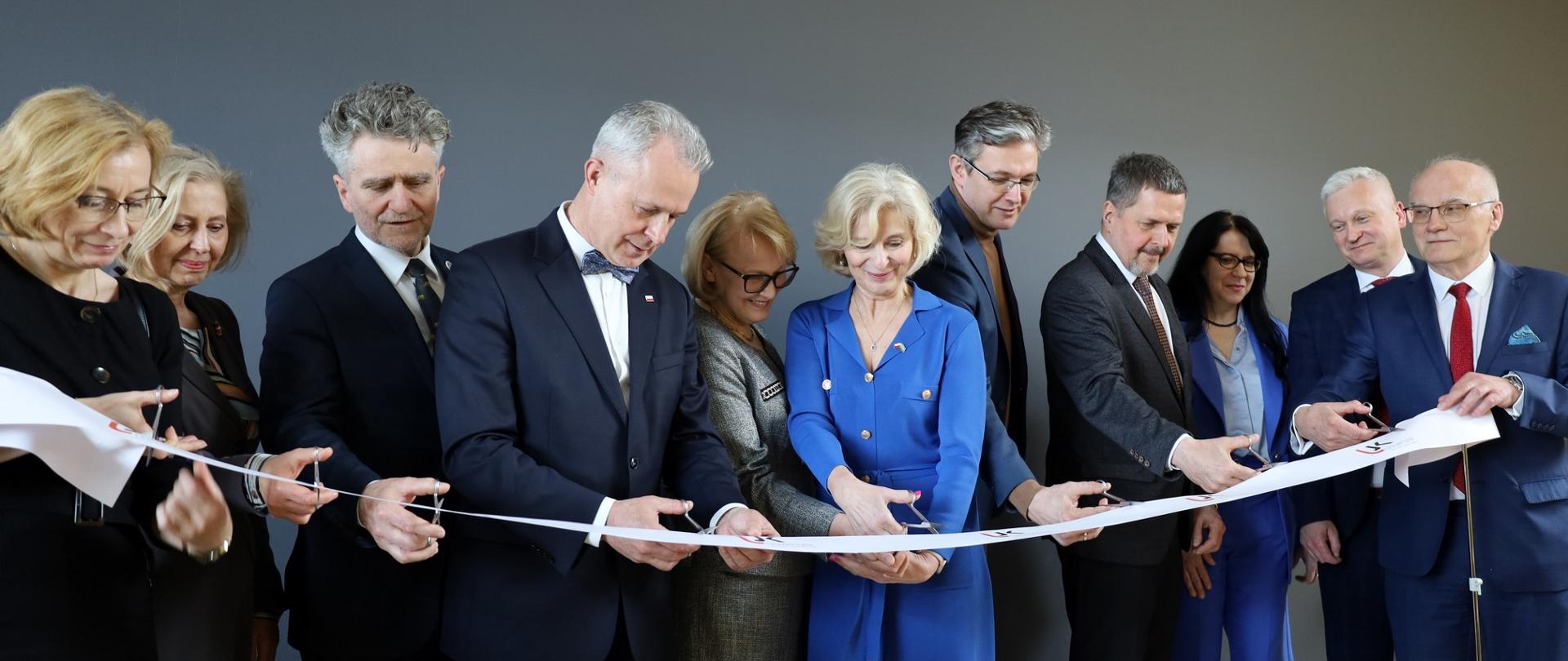 Grupa kilku elegancko ubranych osób stoi przy białej wstędze, każdy ma w ręku nożyczki, w tle ciemnoszara ściana.