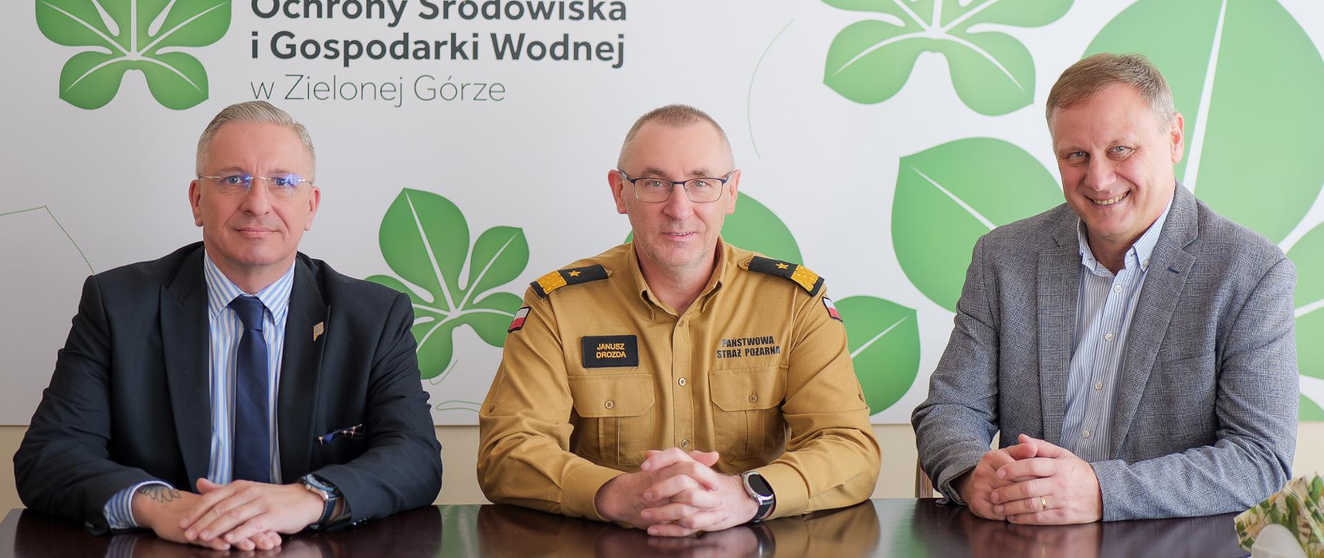 Na zdjęciu przy stole siedzą trzy osoby. W środku znajduje się nadbrygadier Janusz Drozda – Lubuski Komendant Wojewódzki Państwowej Straży Pożarnej, ubrany w mundur koloru beżowego z czarnymi pagonami. Po lewej stronie zdjęcia siedzi Mirosław Glaz – Prezes Zarządu Wojewódzkiego Funduszu Ochrony Środowiska i Gospodarki Wodnej w Zielonej Górze, ubrany w ciemny garnitur i niebieski krawat. Po prawej stronie siedzi Przemysław Nitschka – Zastępca Prezesa Zarządu WFOŚiGW w Zielonej Górze, ubrany w szarą marynarkę i jasną koszulę. W tle widoczne jest logo Wojewódzkiego Funduszu Ochrony Środowiska i Gospodarki Wodnej w Zielonej Górze, zawierające zielone, stylizowane liście na jasnym tle. Wszystkie osoby uśmiechają się lekko, patrząc w kierunku aparatu.
