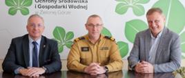 Na zdjęciu przy stole siedzą trzy osoby. W środku znajduje się nadbrygadier Janusz Drozda – Lubuski Komendant Wojewódzki Państwowej Straży Pożarnej, ubrany w mundur koloru beżowego z czarnymi pagonami. Po lewej stronie zdjęcia siedzi Mirosław Glaz – Prezes Zarządu Wojewódzkiego Funduszu Ochrony Środowiska i Gospodarki Wodnej w Zielonej Górze, ubrany w ciemny garnitur i niebieski krawat. Po prawej stronie siedzi Przemysław Nitschka – Zastępca Prezesa Zarządu WFOŚiGW w Zielonej Górze, ubrany w szarą marynarkę i jasną koszulę. W tle widoczne jest logo Wojewódzkiego Funduszu Ochrony Środowiska i Gospodarki Wodnej w Zielonej Górze, zawierające zielone, stylizowane liście na jasnym tle. Wszystkie osoby uśmiechają się lekko, patrząc w kierunku aparatu.