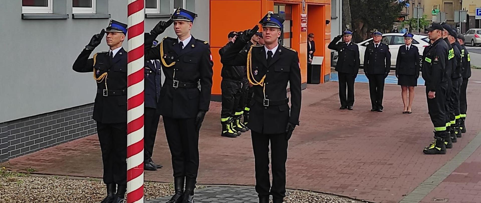 Poczet flagowy salutuje do flagi. W tle strażacy oraz budynek komendy.