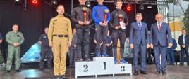 na scenie zawodnicy z pucharami i dyplomami stoją na podium, obok podium straż, i osoby cywilne