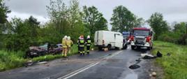 Na zdjęciu widać uszkodzony po czołowym zderzeniu samochód osobowy i dostawczy oraz dwa samochody pożarnicze. Samochody pożarnicze i samochód dostawczy stoją na jezdni, natomiast zniszczony samochód osobowy znajduje się w przydrożnym rowie. Przy pojazdach znajdują się strażacy ubrani w ubrania specjalne i hełmy. 